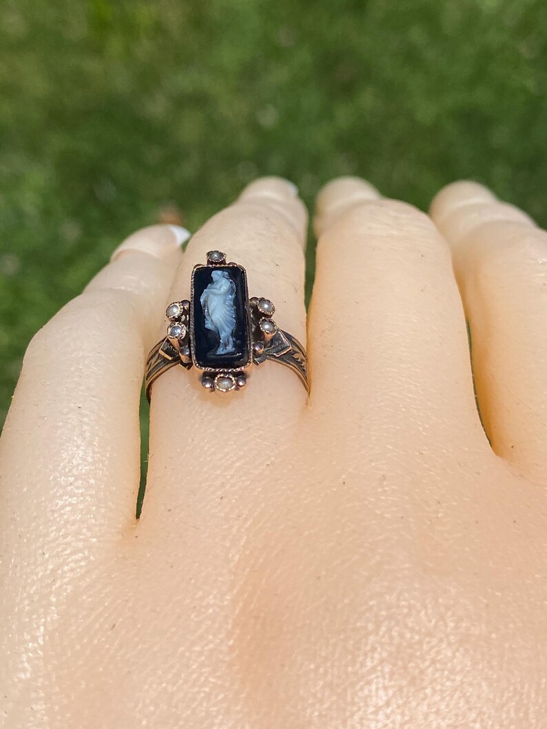 Pu&ograve; includere: Un anello vintage con un cammeo nero rettangolare con una figura bianca. L'anello ha una fascia in metallo color bronzo con accenti decorativi e piccole pietre trasparenti. L'anello &egrave; posto su una mano.