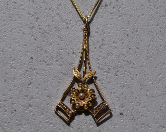 May include: A gold pendant necklace with a delicate floral design. The pendant features a small flower with a pearl center, surrounded by smaller pearls. The pendant is suspended from a delicate gold chain.