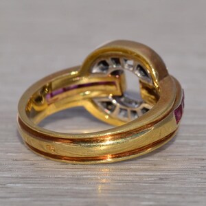 May include: A gold ring with an unusual, interwoven design. The ring has a band with a reddish-brown accent and is decorated with small, red gemstones and clear stones. The ring is set against a light-coloured surface.