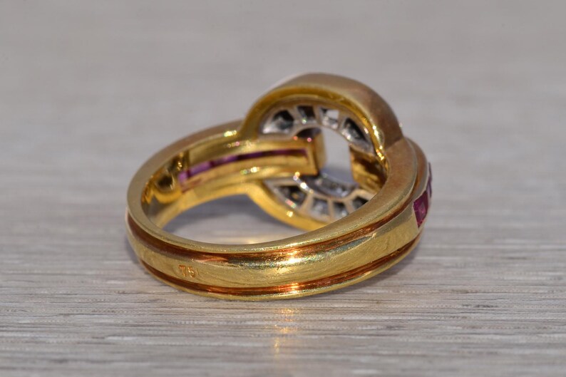 May include: A gold ring with an unusual, interwoven design. The ring has a band with a reddish-brown accent and is decorated with small, red gemstones and clear stones. The ring is set against a light-coloured surface.