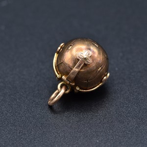 May include: A gold-toned metal ball pendant with a hinged opening and a decorative cross on top. The ball is intricately detailed with a textured surface.