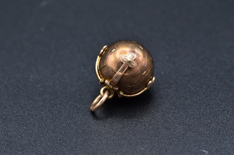 May include: A gold-toned metal ball pendant with a hinged opening and a decorative cross on top. The ball is intricately detailed with a textured surface.