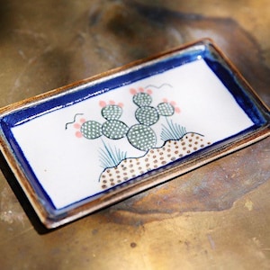 Puede incluir: Bandeja rectangular de cerámica con borde azul y diseño de cactus pintado a mano. El cactus está representado con segmentos verdes punteados, flores rosas y detalles azules. La bandeja tiene un borde marrón y un interior blanco.