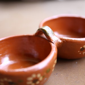 Pode incluir: Um prato duplo de terracota com uma alça que conecta as duas tigelas. As tigelas são decoradas com padrões florais brancos e marrons.