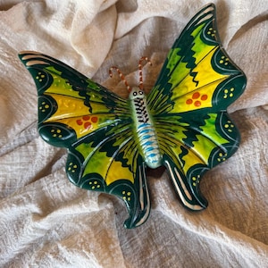 May include: A ceramic butterfly with a vibrant design. The wings are primarily green and yellow, with red and yellow accents. The body is a mix of blue, black, and white. The butterfly is a decorative item.