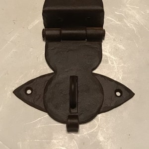 May include: A dark brown metal door hinge with a decorative design. The hinge has a rounded top and a pointed bottom. It has a loop on the bottom for attaching to a door.