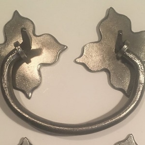 Op de afbeelding: Een set van twee metalen lade-knoppen. Elke knop heeft een decoratieve, bladvormige achterplaat met een gebogen handvat. Het metaal heeft een donkere, verouderde afwerking, wat een vintage of rustieke stijl suggereert. Voor meubels.