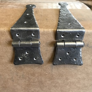 May include: Two black metal hinges with decorative details and multiple holes. The hinges are closed and have a rustic, hammered finish.