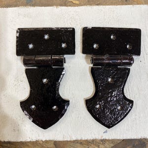 Può includere: Due cerniere decorative nere con finitura strutturata. Ogni cerniera ha una sezione superiore rettangolare con tre fori e una sezione inferiore curva stilizzata con tre fori. Le cerniere sono collegate da un giunto cilindrico.