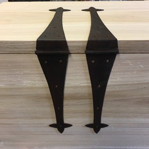 Puede incluir: Dos bisagras de metal negro con extremos puntiagudos decorativos. Las bisagras están unidas a una superficie de madera clara.