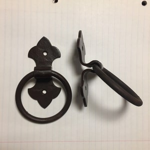 Puede incluir: Dos tiradores de puerta de metal marrón oscuro de estilo antiguo. Uno es un tirador de anillo con una placa trasera decorativa, y el otro es un tirador de bisagra a juego. El herraje está diseñado para una estética rústica o vintage.