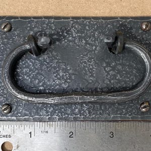 May include: A dark gray, rectangular drawer pull with a hammered texture and a curved handle. The pull is attached with four screws. A ruler shows the pull is approximately 4 inches long.