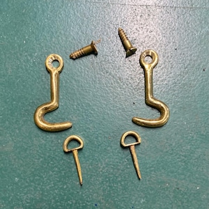 May include: A collection of brass hardware components, including two J-shaped hooks, two small D-ring pins, and two wood screws. The hardware is displayed against a green surface, showcasing the metallic sheen of the brass.