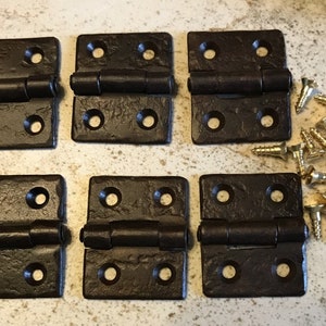 May include: Six dark brown metal hinges with a hammered finish and a set of gold screws. The hinges are square shaped with rounded corners and have four holes on each side.