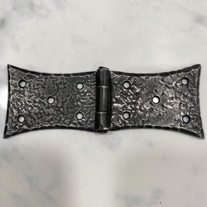 May include: A dark gray metal strap hinge with a textured, hammered surface finish. The hinge features a central barrel joint connecting two symmetrical, flared wings, each containing four circular screw holes for mounting purposes.