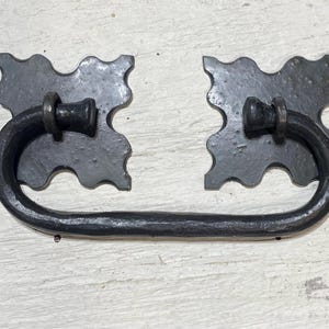 Könnte beinhalten: Schwarzer Metall-Schubladengriff mit dekorativem, gebogenem Griff. Der Griff hat zwei quadratische Rückplatten mit einem gewellten Randdesign. Der Griff hat eine halbkreisförmige Form und die Hardware hat ein rustikales, antikes Aussehen.