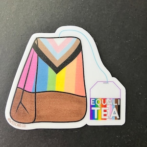 Può includere: Un adesivo di una bustina di tè con strisce arcobaleno e marroni, con un'etichetta che dice "EQUALI TEA" in colori arcobaleno.