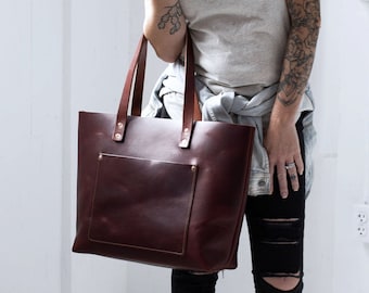May include: A brown leather tote bag with a front pocket. The bag has a long strap and is worn by a person wearing a grey t-shirt and ripped jeans.