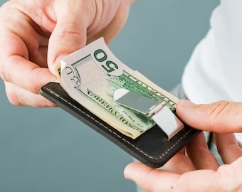 May include: A brown leather money clip with a silver metal money clip holding a stack of US dollar bills. The bill on top is a $50 bill.