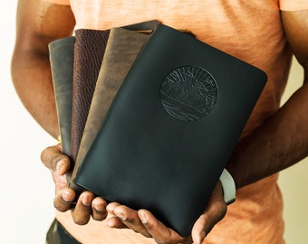 May include: A stack of four leather-bound journals, the top one is black with an embossed design of a mountain range and a sun. The journals are held in a person's hands.