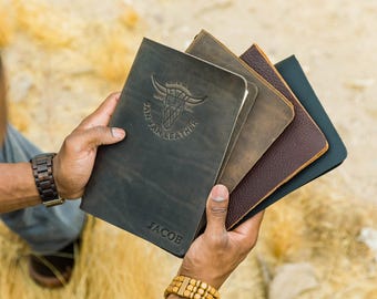 Può includere: Una collezione di quattro diari in pelle in varie tonalità di marrone, grigio e nero. Il diario frontale presenta un logo con teschio di toro in rilievo con il testo SAN TAN LEATHER e il nome JACOB impresso sul bordo inferiore della copertina.