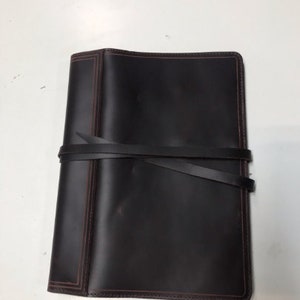 May include: A dark brown leather notebook cover with a strap closure and red stitching. The cover has a worn, distressed look.