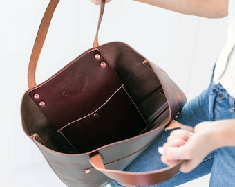 May include: A brown leather tote bag with a large pocket on the inside. The bag has a brown leather strap and a logo that says "Sam & Sons Leather" on the front.