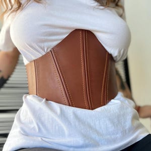 May include: A brown corset with vertical seams and yellow stitching. The corset is worn over a white t-shirt. The corset is made of a smooth material and has a structured design.