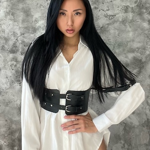 May include: A woman wearing a white button-down shirt with a black leather belt with silver buckles. The belt has two straps that go over the shoulders.