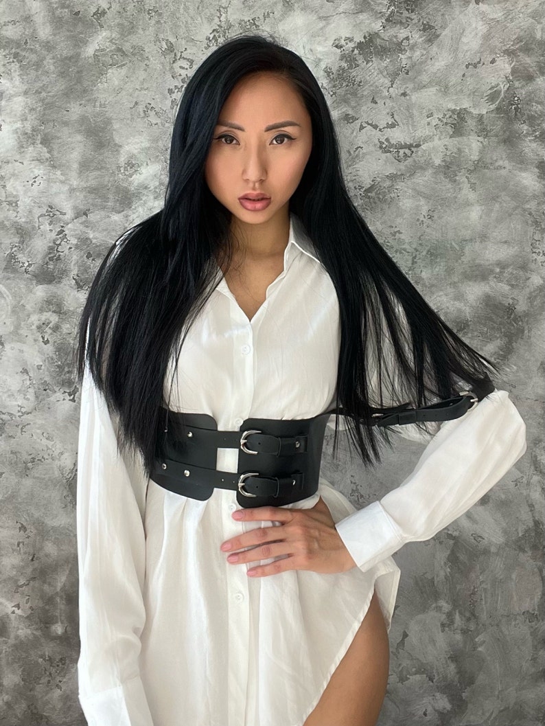 May include: A woman wearing a white button-down shirt with a black leather belt with silver buckles. The belt has two straps that go over the shoulders.