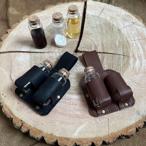 Może przedstawiać: Małe szklane butelki z korkami, niektóre wypełnione płynami i proszkami. Pokazano dwa czarne skórzane uchwyty, każdy mieszczący dwie butelki, oraz brązowy skórzany uchwyt. Przedmioty ułożone są na drewnianej powierzchni.