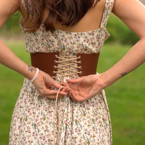 May include: A brown corset cinches the waist of a floral print dress. The dress features a cream background with small blue, red, and green flowers. The corset is laced up with a light-colored cord, creating a vintage aesthetic.