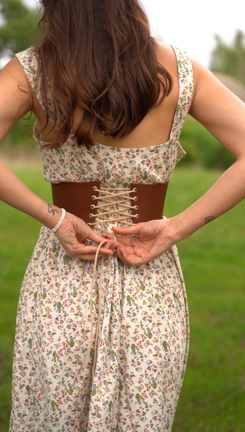 May include: A brown corset cinches the waist of a floral print dress. The dress features a cream background with small blue, red, and green flowers. The corset is laced up with a light-colored cord, creating a vintage aesthetic.