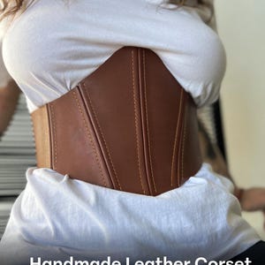 May include: A handmade brown leather corset with visible stitching. The corset is worn over a white shirt. The text "Handmade Leather Corset" and "Premium Craftsmanship and Customizable Details" are displayed at the bottom of the image.