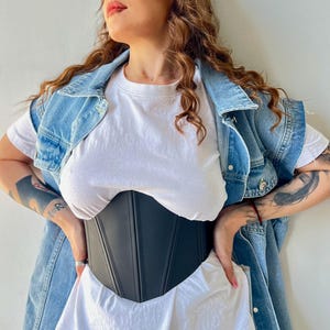 May include: A black corset with vertical seams is worn over a white t-shirt. A denim vest is worn over the t-shirt. The model is wearing black leggings. The corset is a waist-cinching garment.