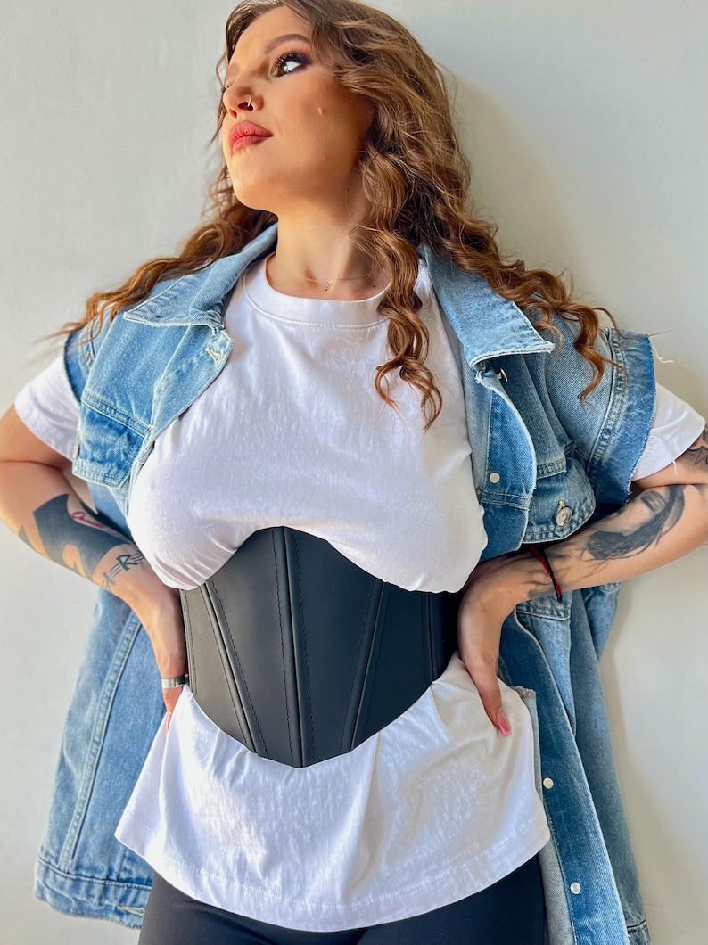 May include: A black corset with vertical seams is worn over a white t-shirt. A denim vest is worn over the t-shirt. The model is wearing black leggings. The corset is a waist-cinching garment.