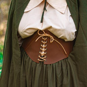 Può includere: Un corsetto in pelle marrone con lacci color crema è il punto focale dell'immagine. È indossato sopra una camicetta color crema e una gonna verde scuro. Un mantello verde scuro è drappeggiato sulle spalle.