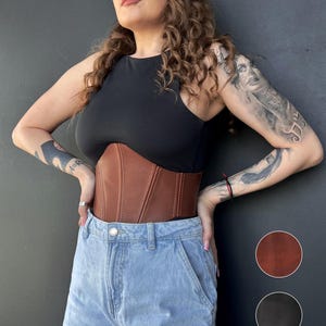 May include: A brown leather corset with vertical stitching. The corset is worn over a black top and paired with light blue denim jeans. Two color swatches, brown and black, are shown on the right side of the image.