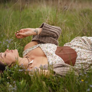 May include: A person wearing a floral print dress and brown corset lies in a field of green grass and wildflowers. The person is also wearing a brown sweater. The scene evokes a sense of peace and tranquility.