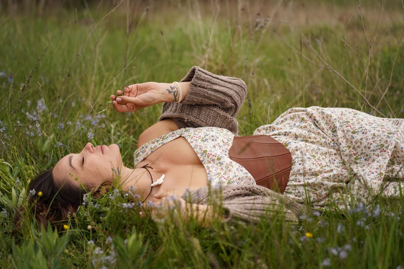 May include: A person wearing a floral print dress and brown corset lies in a field of green grass and wildflowers. The person is also wearing a brown sweater. The scene evokes a sense of peace and tranquility.