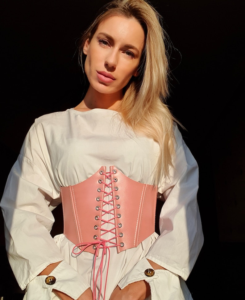 May include: A pink leather corset belt with a lace-up front. The belt is tied in a bow at the front and has silver eyelets.