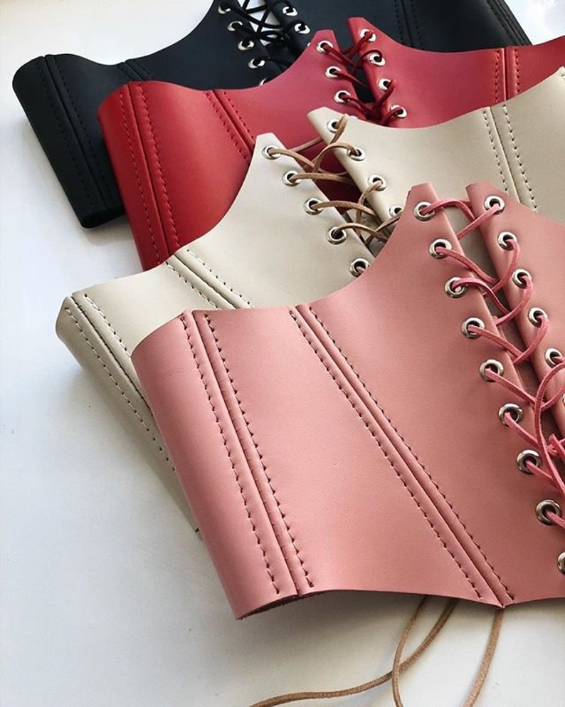May include: Four different colored leather corsets with lace-up fronts. The corsets are stacked on top of each other, with the pink corset on the bottom.