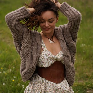 May include: A woman wearing a brown cable-knit cardigan over a floral print dress with a brown leather corset. The dress has a white background with small colorful flowers. A necklace with a white pendant is visible.