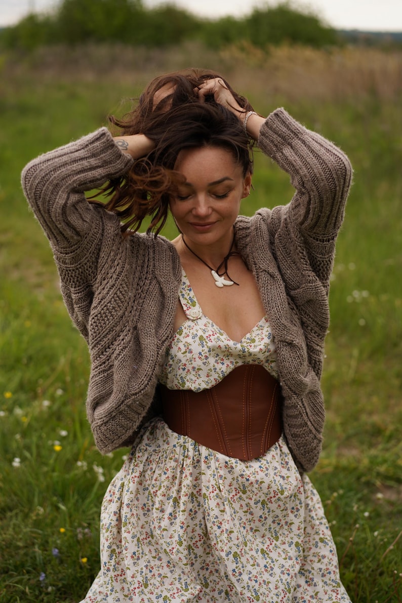 May include: A woman wearing a brown cable-knit cardigan over a floral print dress with a brown leather corset. The dress has a white background with small colorful flowers. A necklace with a white pendant is visible.