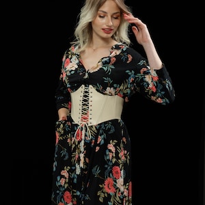 May include: A woman wearing a black floral dress with a white corset belt. The dress has a long sleeve and a v-neckline. The corset belt has a lace-up front.