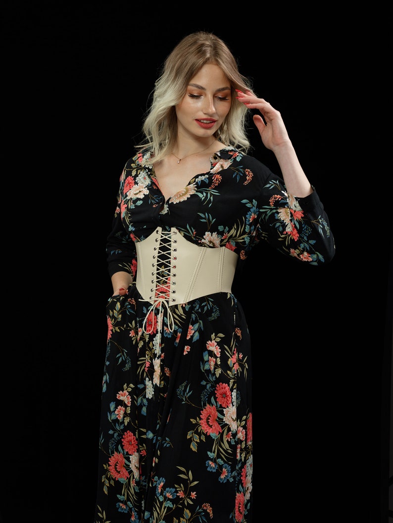 May include: A woman wearing a black floral dress with a white corset belt. The dress has a long sleeve and a v-neckline. The corset belt has a lace-up front.