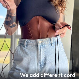 May include: A brown leather corset with a black top and light blue jeans. The corset has vertical stitching and a cinched waist. The image includes the text: "We add different cord options in the kit!"