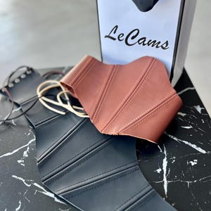 May include: Two corsets, one black and one brown, are displayed on a black and white marble surface. The brown corset is on top of the black one. A white shopping bag with the text "Le Cams" is in the background.