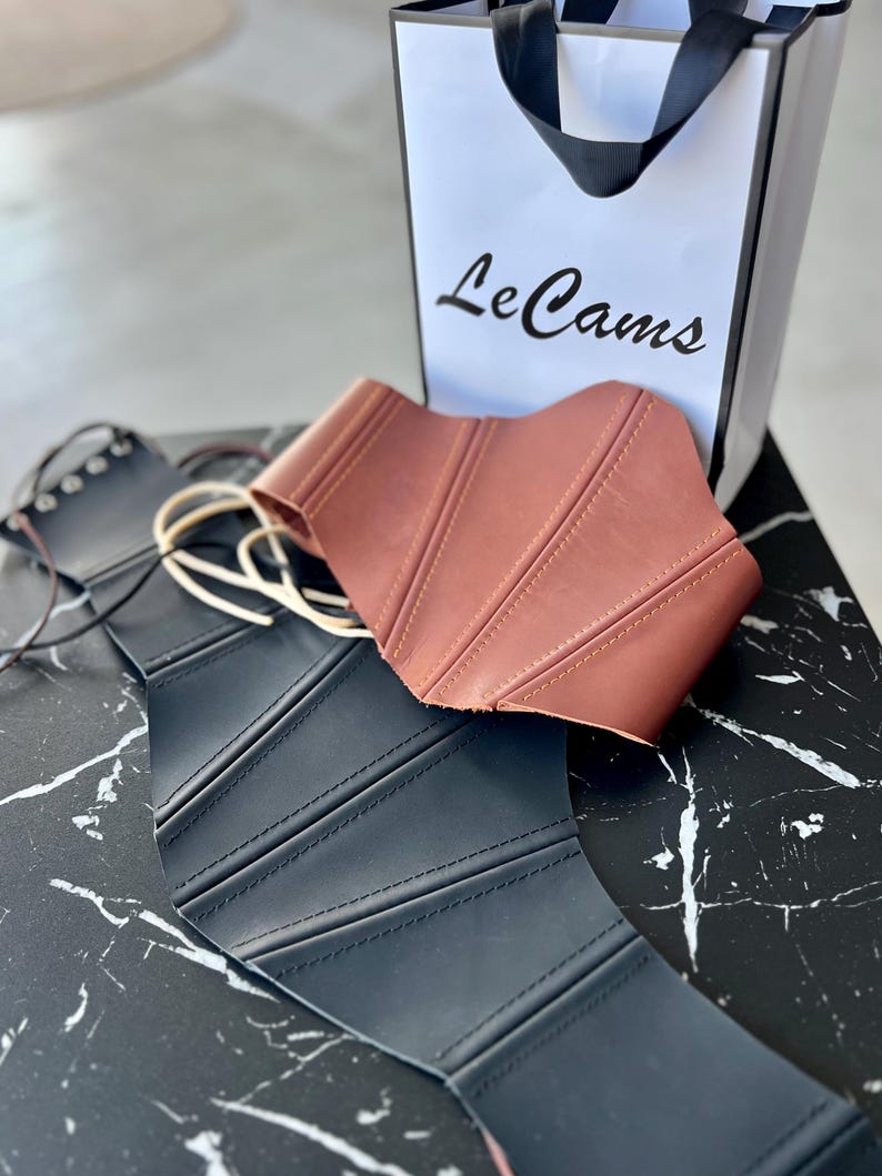 May include: Two corsets, one black and one brown, are displayed on a black and white marble surface. The brown corset is on top of the black one. A white shopping bag with the text "Le Cams" is in the background.