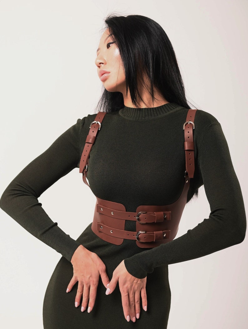 May include: A brown leather harness with double buckles and adjustable straps. The harness is worn over a dark green long-sleeved shirt.
